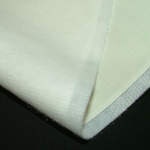 Puede incluir: Tela blanca con una textura suave y peluda en un lado y una textura lisa y tejida en el otro lado. La tela tiene un borde blanco cosido.