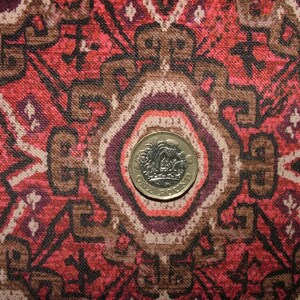 May include: Close-up of a patterned fabric with a red, brown, and cream color scheme. The design features geometric shapes and floral motifs. A gold-colored one-pound coin is centered on the fabric, providing a size reference.