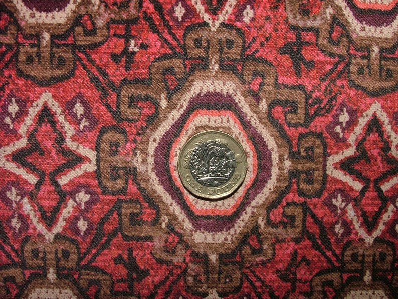 May include: Close-up of a patterned fabric with a red, brown, and cream color scheme. The design features geometric shapes and floral motifs. A gold-colored one-pound coin is centered on the fabric, providing a size reference.