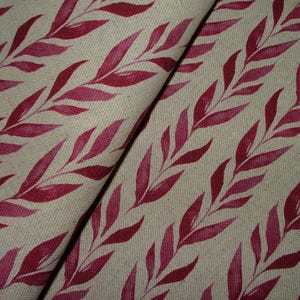 May include: A close-up of beige fabric featuring a repeating pattern of pink and red leaves. The leaves are painted in a watercolor style and arranged diagonally. The fabric has a textured appearance, creating a natural design.