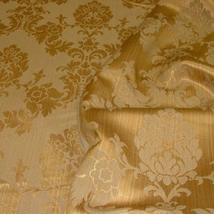K&ouml;nnte beinhalten: Nahaufnahme eines goldenen Stoffes mit Damastmuster. Der Stoff hat einen dezenten Glanz und ein erhabenes Design.