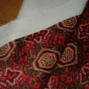 May include: Close-up of two fabrics. One is a white, textured fabric with a raw edge and a stitched seam. The other is a patterned fabric with a red background and brown and orange geometric designs.