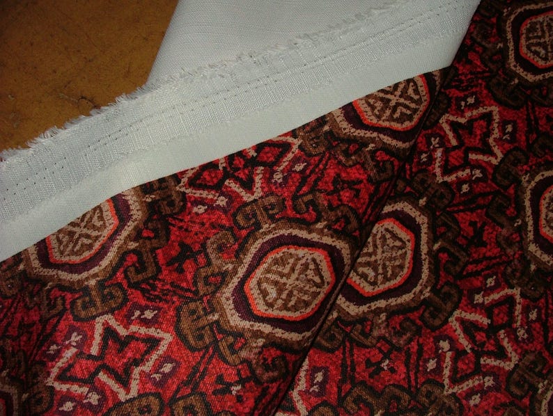 May include: Close-up of two fabrics. One is a white, textured fabric with a raw edge and a stitched seam. The other is a patterned fabric with a red background and brown and orange geometric designs.