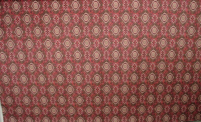 May include: A patterned fabric with a repeating design of diamond and circular shapes in shades of red, brown, and beige. The fabric has a textured appearance, creating a visually rich surface.