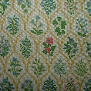 May include: A close-up of a fabric with a repeating floral pattern. The design features various flowers in shades of blue, green, and pink, set against a cream-coloured background. A gold-coloured trellis-like pattern frames the floral elements.