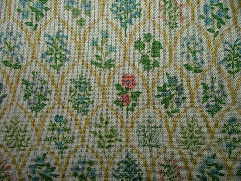 May include: A close-up of a fabric with a repeating floral pattern. The design features various flowers in shades of blue, green, and pink, set against a cream-coloured background. A gold-coloured trellis-like pattern frames the floral elements.