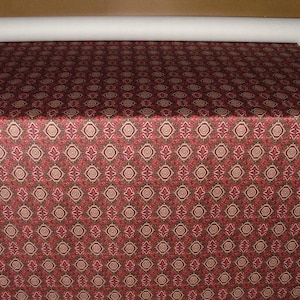 May include: A roll of fabric with a repeating pattern of red, brown, and beige geometric shapes. The fabric is spread out, showing the detailed design. A roll of white fabric is visible at the top.