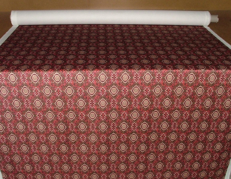 May include: A roll of fabric with a repeating pattern of red, brown, and beige geometric shapes. The fabric is spread out, showing the detailed design. A roll of white fabric is visible at the top.