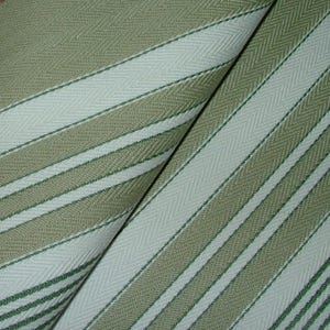 May include: Close-up of a folded fabric with a striped pattern. The fabric features alternating stripes of light green, off-white, and darker green. The stripes run diagonally across the image, creating a textured appearance. The fabric appears to be woven.