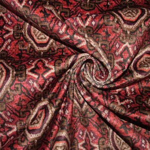 May include: Close-up of a patterned fabric with a swirling design. The fabric features a repeating pattern of geometric shapes in shades of red, brown, and cream. The fabric is folded and draped, showing the texture and detail of the design.
