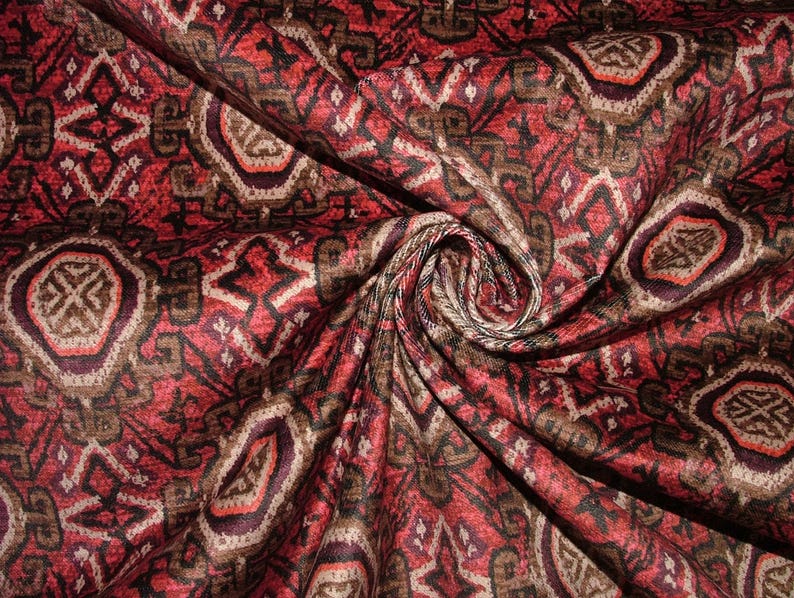 May include: Close-up of a patterned fabric with a swirling design. The fabric features a repeating pattern of geometric shapes in shades of red, brown, and cream. The fabric is folded and draped, showing the texture and detail of the design.
