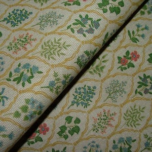 May include: Close-up of fabric with a repeating floral pattern. The off-white fabric features a grid of yellow outlines, each containing a different floral design in shades of green, pink, and blue. The fabric is folded, showing its texture.