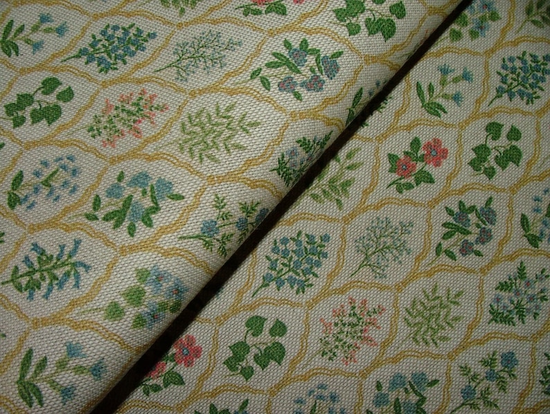 May include: Close-up of fabric with a repeating floral pattern. The off-white fabric features a grid of yellow outlines, each containing a different floral design in shades of green, pink, and blue. The fabric is folded, showing its texture.