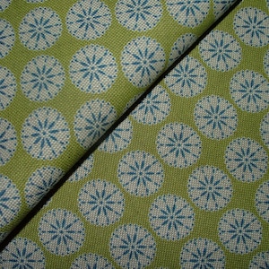 May include: Close-up of a green fabric with a repeating pattern of white and teal circular designs. The fabric is folded, showing the texture and the detailed pattern. The design is reminiscent of stylized flowers or starbursts.