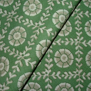 Pode incluir: Close-up de tecido verde com um padrão floral repetitivo. O design apresenta flores brancas e bege estilizadas e ramos com folhas. O tecido está dobrado, mostrando a textura e profundidade do material.