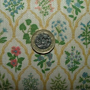 May include: A close-up of a fabric featuring a floral pattern in blue, pink, and green on a cream background. A one-pound coin is placed on the fabric, providing a size reference. The fabric has a repeating pattern of flowers and leaves.