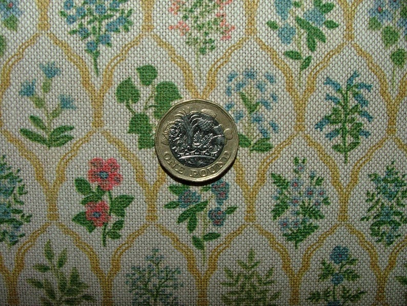 May include: A close-up of a fabric featuring a floral pattern in blue, pink, and green on a cream background. A one-pound coin is placed on the fabric, providing a size reference. The fabric has a repeating pattern of flowers and leaves.