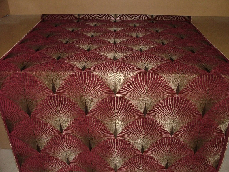 May include: Red fabric with a gold geometric pattern of overlapping fans. The fabric has a subtle sheen and a textured surface.