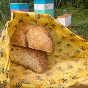 May include: A yellow beeswax food wrap with a honeycomb and bee pattern is used to wrap two slices of sourdough bread.