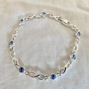 May include: A silver bracelet featuring an infinity symbol design, interspersed with round, blue gemstones. The bracelet has a clasp closure and is displayed on a white surface. A delicate piece of jewelry.