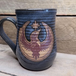 May include: A blue ceramic mug with a handle, featuring a detailed emblem of a bird with outstretched wings and a star. The bird is brown and gold, set against a blue background. The mug has a textured surface.