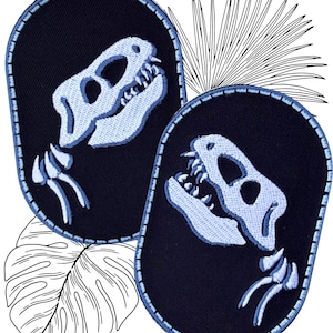 Op de afbeelding: Twee zwarte ovale patches met witte geborduurde dinosaurus skeletten. De dinosaurus skeletten zijn naar rechts gericht en hebben een gedetailleerde schedel en kaak.