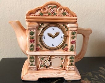 May include: A ceramic teapot shaped like a grandfather clock with a white face and black hands. The teapot is decorated with floral details and has a gold trim.
