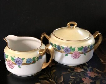 May include: A vintage porcelain creamer and sugar bowl set with a floral design. The set features a yellow background with a band of purple, pink, and blue flowers with green leaves. The handles are gold.