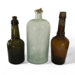 May include: Three antique glass bottles with cork stoppers. The bottles vary in color, including green, clear, and amber. The bottles are different shapes and sizes, suggesting they may have been used for different purposes.