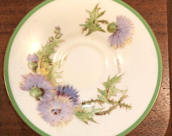 May include: A white saucer with a green rim features a hand-painted design of purple and green thistles. The saucer is marked "England" in the center.