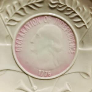 May include: A white ceramic plate with a pink and white medallion in the center. The medallion features a profile of a person and the text "Declaration of Independence 1776".