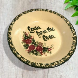 May include: A cream-colored pie plate with a green rim and a floral design. The plate has the words "Lovin' from the Oven" written on it.