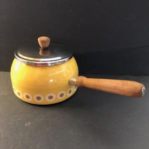 May include: Yellow enamel saucepan with a stainless steel lid and wooden handle. The saucepan has a decorative floral pattern around the base. The lid has a wooden knob. A vintage kitchenware item.