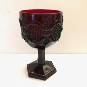 May include: A deep red glass goblet featuring a textured diamond pattern on the bowl, accented by raised circular motifs. The stem connects to a hexagonal base with a ridged edge, creating a structured silhouette for this pressed glass vessel.