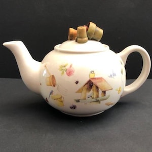 May include: Cream-colored ceramic teapot with a lid and handle. The teapot is decorated with a birdhouse, flowers, and small buckets. The lid is topped with three small, decorative flower pots.