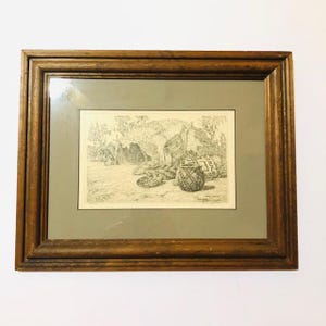 May include: A framed black and white print depicting a rural scene with a dwelling, baskets, and other objects. The artwork is set against a gray mat and encased in a dark brown wooden frame. The image has a vintage aesthetic.