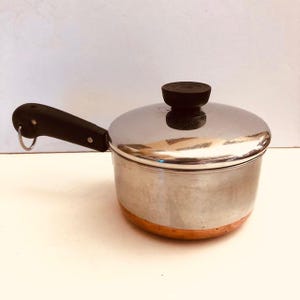May include: A stainless steel saucepan with a copper-colored base and a black handle. The pot has a matching lid with a black knob. The handle has a metal ring attached. The pot is designed for cooking.
