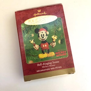 May include: A Hallmark Christmas ornament featuring Mickey Mouse dressed as Santa Claus, holding golden bells. The ornament is in a decorative box with the text "Bell-Ringing Santa" and "Mickey Mouse". The box has a red and gold color scheme.