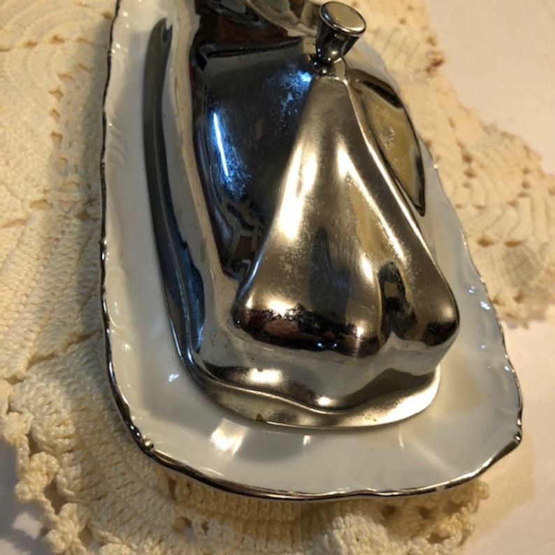 May include: A silver butter dish with a white porcelain base and a scalloped edge. The lid has a knob on top.