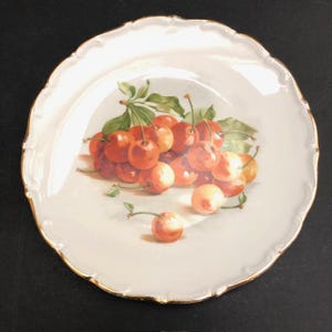 May include: A white porcelain plate with a scalloped edge and gold trim. The plate features a detailed painting of ripe cherries with green leaves. The cherries are a mix of red and orange hues, creating a vibrant display.