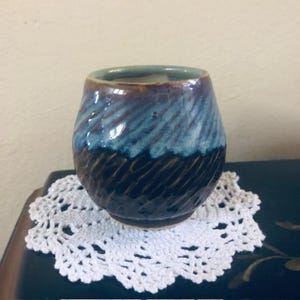 May include: A handmade ceramic tumbler with a textured surface. The top is a teal green, transitioning to a blue and brown swirl pattern. The bottom half is a dark brown with carved lines. The tumbler sits on a white lace doily.