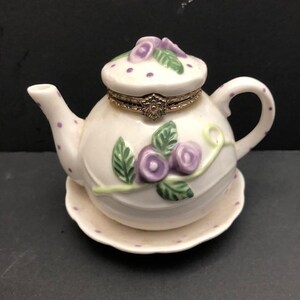 May include: A white ceramic teapot-shaped trinket box with a removable lid. The surface features purple polka dots, sculpted green leaves, and purple floral accents. A gold-toned metal hinge with a decorative floral clasp connects the lid.