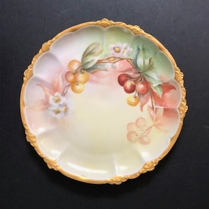 May include: A vintage porcelain plate with a scalloped edge and gold trim. The plate features a hand-painted design of cherries and flowers in shades of pink, yellow, and green.