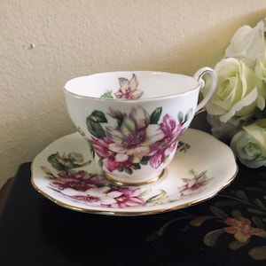 May include: A white teacup and saucer with a floral pattern. The flowers are pink, white, and green. The cup and saucer have a gold trim.