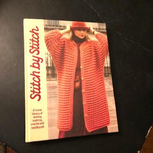 May include: A book titled "Stitch by Stitch" with a home library of sewing, knitting, crochet and needle craft. The cover features a woman wearing a red knitted cardigan.