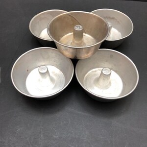 May include: A collection of five vintage aluminum angel food cake pans. Each pan has a central tube and a rounded bowl shape. The pans are arranged on a dark surface, showcasing their metallic silver color and classic design.