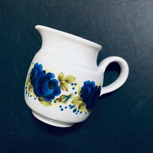 May include: White ceramic creamer with a blue and green floral design. The creamer has a rounded body and a handle.