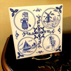 May include: A white ceramic tile with a blue and white design featuring four scenes of Dutch life: a woman carrying a basket of flowers, a windmill, a sailboat, and a man carrying a bundle of reeds. The tile is on a decorative stand.