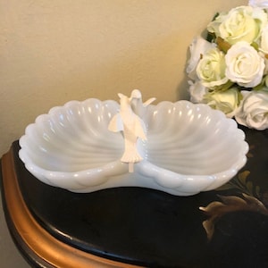 May include: A white ceramic double dish with a bird figurine handle. The dish is shaped like a seashell and has a scalloped edge.