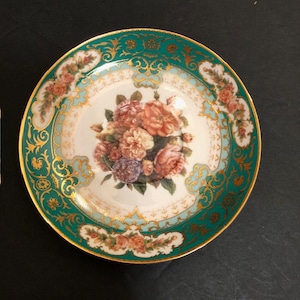 May include: A white porcelain saucer with a green and gold rim. The center of the saucer features a bouquet of pink and white roses with green leaves.